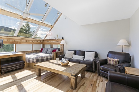 A sunlit room with a sloped glass ceiling, featuring a bed, two dark leather chairs, a wooden coffee table, and a scenic view through large windows.