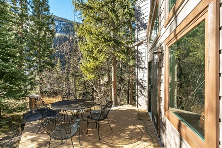 Wooden deck with a round table and four chairs, adjacent to a house, surrounded by forested trees and mountains in the background on a sunny day.