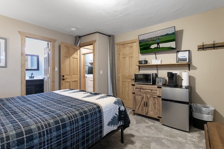 A small bedroom with a bed, dresser, TV, microwave, mini fridge, and coffee maker. A folding door opens to a closet, and an open door reveals part of a bathroom. Walls are beige with wooden trim.