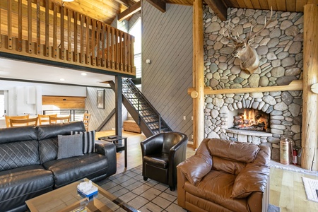 Living room with stone fireplace, mounted deer head, leather chairs and couches, wooden ceiling beams, staircase, and a mezzanine with railing.