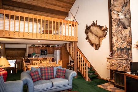 A cozy living room features a stone fireplace, mounted animal decor, and a lofted bedroom area. The wood-paneled kitchen and dining set are visible, with a blue sofa and plaid pillows in the foreground.