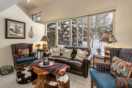 Cozy living room with large windows showing a snowy outdoor scene. The room features a leather sofa with several pillows, plaid armchairs, a rustic wooden coffee table, and various decorations.