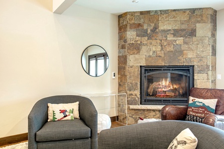 A cozy living room with a stone fireplace, two gray armchairs, a leather chair, and a glass side table. A round mirror and decorative pillows are placed around the space.