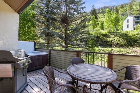 A wooden deck with a round table and four chairs, a gas grill, and a hot tub, overlooking a wooded hillside on a sunny day.