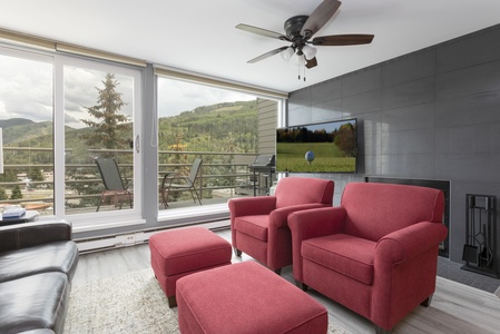 Living room with red armchairs, matching ottomans, and a black leather sofa. A large window overlooks a scenic view of hills. A ceiling fan and wall-mounted TV are present.