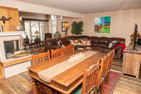 A living room with a large brown leather sectional sofa, a wooden dining table with chairs, a fireplace, and a television. A painting hangs on the wall, and a rug is under the dining table.