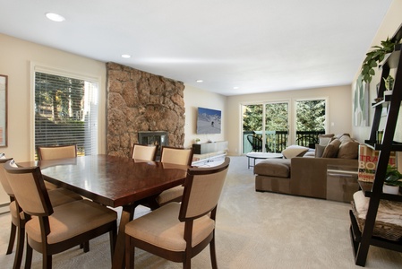 A spacious living room with a dining table, chairs, a brown sectional sofa, a stone fireplace, and large windows with a view of greenery.