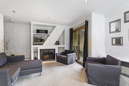 A living room with black armchairs, a fireplace, a TV mounted above it, white walls, geometric shelving, and glass doors leading to an outdoor space.