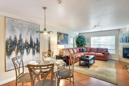 A cozy living room with a brown leather sectional sofa, a dining table set for four, and contemporary art on the walls. The room has wooden flooring and large windows allowing natural light.