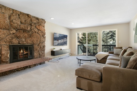 A cozy living room with a large stone fireplace, a sectional sofa, a round coffee table, and a large window with a view of trees. A painting hangs above the sofa.