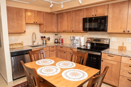 A modern kitchen with wooden cabinets, stainless steel appliances, and a countertop with small kitchen appliances. A wooden dining table with four chairs and decorative placemats is in the center.