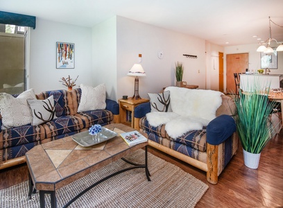 A cozy living room with two patterned sofas, a wooden coffee table with a magazine, a lamp on a side table, and a plant. The room leads into a dining area with a hanging light fixture.