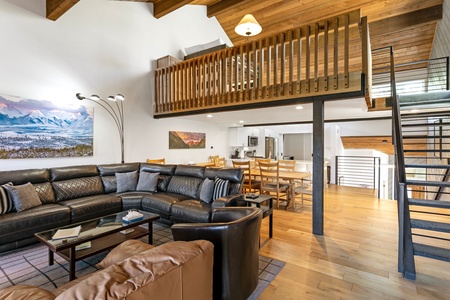 Spacious living area with black leather sofas, a brown leather chair, wooden floors, a loft space, a staircase to the right, and a dining area adjacent to the kitchen in the background.