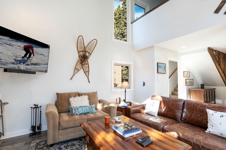 A cozy living room with a brown leather sofa, a beige loveseat, a wooden coffee table, and a TV displaying a skiing scene. Snowshoes are mounted on the wall, and a staircase is visible in the background.