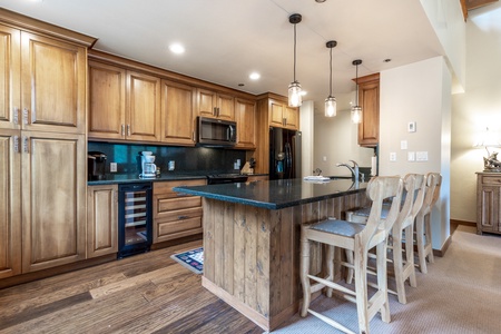 A modern kitchen with wooden cabinets, an island with seating, black countertops, and stainless steel appliances under pendant lights.