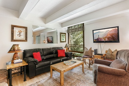 A living room with a black leather sofa, a brown armchair, and a wooden coffee table. Wall decor includes a large mirror and a framed painting. A TV is mounted on the wall.