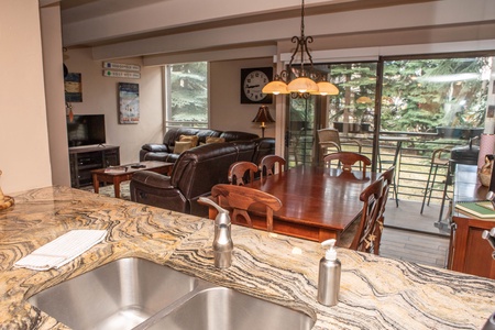 Open living and dining area with a marble countertop in the foreground, sofa and chairs around a dining table, TV, and large window with a sliding door leading to a balcony with outdoor greenery.