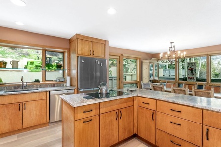 A spacious kitchen and dining area with wooden cabinets, granite countertops, stainless steel appliances, and large windows providing ample natural light. A chandelier hangs above the dining table.