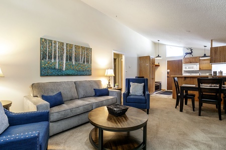 Living room with a gray sofa, blue armchairs, a round coffee table, wall art, and a dining area with a table and chairs. Kitchen in the background with wooden cabinets and white appliances.