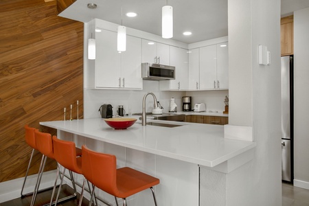 Modern kitchen with white cabinetry, stainless steel appliances, and a white countertop island. Features three orange barstools and wooden accent wall.