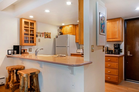 A small kitchen with wooden cabinets, a refrigerator, and a countertop with two wooden stools. There are light fixtures on the ceiling and a piece of artwork on the wall.