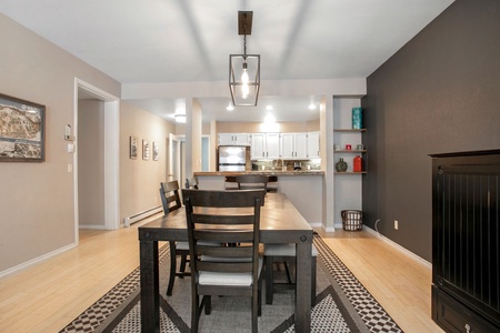 Modern dining area with a rectangular wooden table and six chairs, pendant light above, and open kitchen in the background. Walls are painted in gray tones with artwork and shelves for decoration.