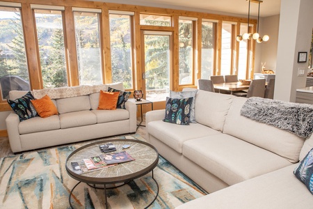 A modern living room with large windows, two beige sofas with patterned cushions, a round coffee table with magazines, and a dining area in the background with a table and chairs.