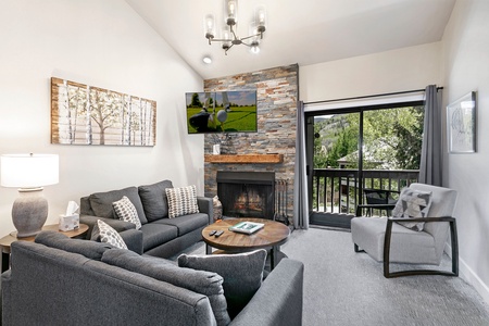 A modern living room features a grey sectional sofa, a single grey armchair, a round coffee table, a stone fireplace, wall-mounted TV, framed artwork, and sliding glass doors leading to a balcony.