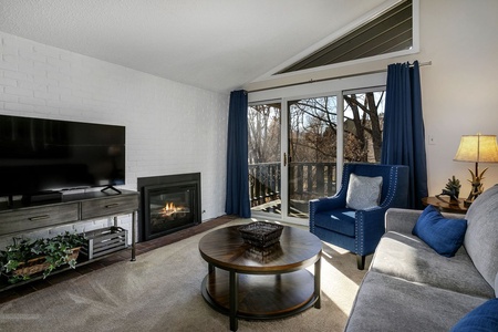 A living room with a gray sofa, blue armchair, round wooden coffee table, fireplace, flat-screen TV, and sliding glass doors with blue curtains leading to a balcony.