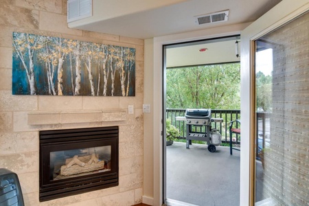 A living room with a stone fireplace and painting above it. A glass door opens to a balcony with a grill and greenery outside.