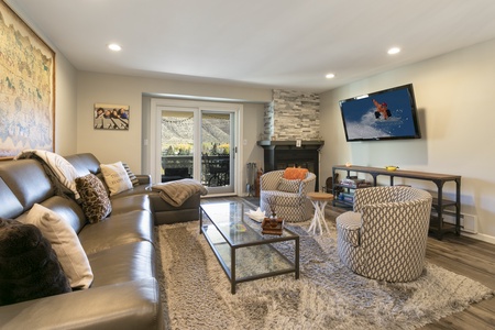 A cozy living room with a large sofa, round chairs, a glass coffee table, a fireplace, and a wall-mounted TV. A sliding door opens to a balcony with a mountain view.