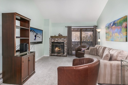 A living room with a fireplace, flat-screen TV on the wall, large brown sofa, brown chair, and a wooden cabinet. A colorful painting is on the wall, and a glass door leads to an outdoor area with trees.