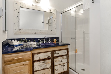 A compact bathroom with a blue-tiled countertop, wooden drawers, a large mirror, and a glass-enclosed shower.