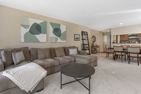 A modern living room features a gray sectional sofa, abstract wall art, a round coffee table, and a dining area with a wooden table and chairs in the background.