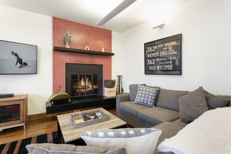 A cozy living room with a lit fireplace, a gray couch, and a coffee table. Shelves with decor and a framed map hang on the walls. A large TV is mounted beside the fireplace.