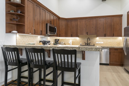 A modern kitchen with wooden cabinets, marble countertops, a stainless steel refrigerator, a stove, a microwave, and three black stools at a breakfast bar.