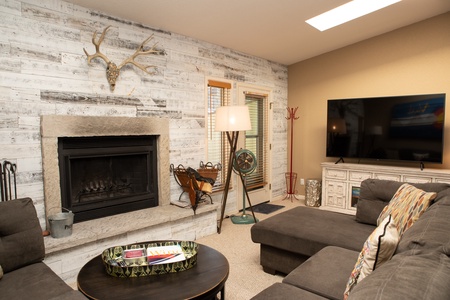 A cozy living room with a stone fireplace, a wall-mounted antler decor, a large TV, a sectional sofa, and a coffee table with magazines. The room features light wood and neutral color tones.