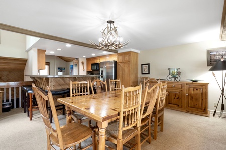 A dining room with a wooden table and eight chairs, an antler chandelier, a sideboard, and an open view to a kitchen with a bar counter and stainless steel appliances.