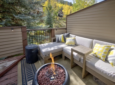 A cozy outdoor patio with a sectional sofa, colorful pillows, a wooden side table, and a fire pit with a flame. The area is surrounded by trees and greenery.