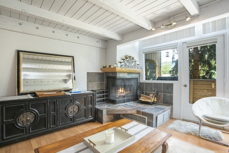 A cozy living room with a modern fireplace, white ceiling beams, large mirror, dark cabinet, sliding glass doors, and a wooden coffee table with a candle. The décor is minimalist and bright.