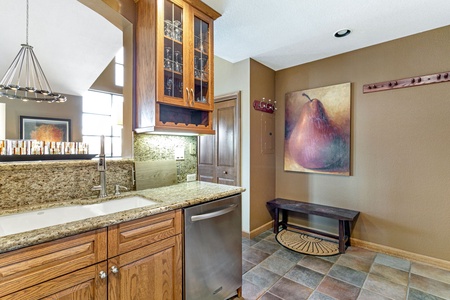 A kitchen area with wooden cabinets, granite countertops, a stainless steel dishwasher, and a small adjacent hallway displaying a large pear painting and a bench.