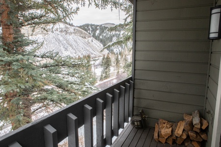 A small covered balcony with a stack of firewood overlooks snowy mountains and a pine tree. The balcony railing is painted dark gray.