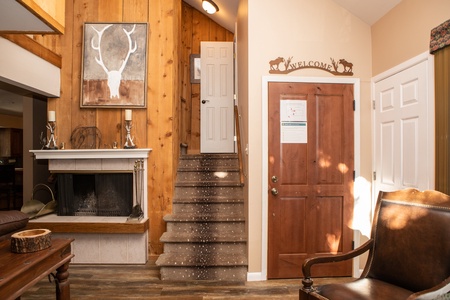 A rustic interior features a wooden-paneled wall, deer antler artwork, a fireplace, a staircase leading upstairs, and a “Welcome” sign above a wooden door. A brown leather chair is placed nearby.