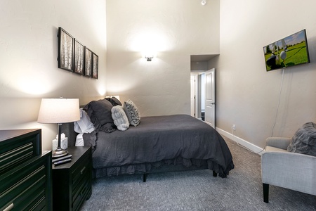 A bedroom with a gray bed, nightstand with lamp, wall-mounted TV, artwork above the bed, and door leading to an adjoining room.