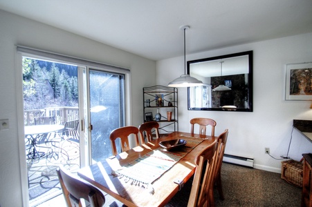 A dining room with a wooden table and chairs, pendant light, large wall mirror, and a sliding glass door leading to an outdoor deck with patio furniture.