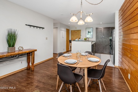 A dining area with a round wooden table set for four, black chairs, and a potted plant on a console table against the wall. The adjacent kitchen features a counter, stainless steel refrigerator, and wooden accents.