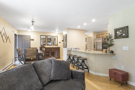 Open living area with a gray sofa, bar stools at a granite kitchen counter, dining table with chairs, and modern decor in a well-lit space.