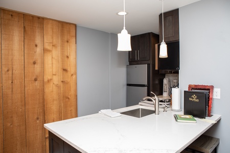 A modern kitchen with a marble countertop, a sink, a stainless steel refrigerator, wooden cabinets, and two hanging pendant lights. A small stack of items is placed on the counter next to the sink.