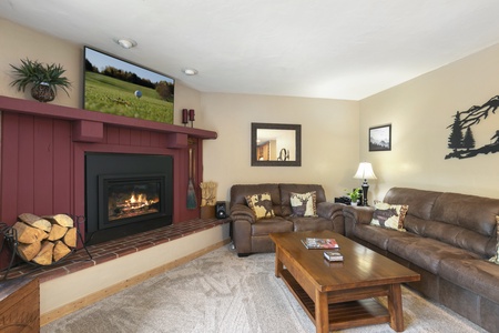 A cozy living room with a fireplace, brown leather sofas, a wooden coffee table, a TV mounted above the mantel, and decorative wall art. A stack of firewood is beside the fireplace.