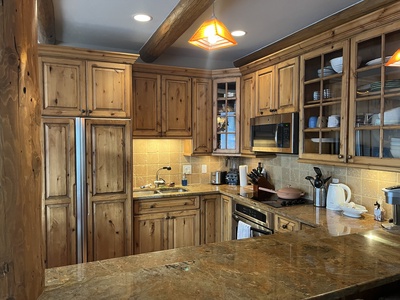 A kitchen with wooden cabinets, a stainless steel fridge, a sink, a microwave, an oven, and various kitchen utensils. The countertop is made of marble and there are two pendant lights overhead.